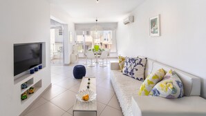A modern living room with a flat-screen TV, a coffee table with decorative items, a sofa with patterned cushions, and a dining area in the background. - Protaras Pallini Seaside Suite (Protaras)