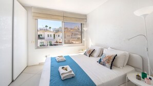 A bedroom with a large bed, a window with a view of buildings, a bedside table with a lamp, and a nightstand. - Protaras Pallini Seaside Suite (Protaras)