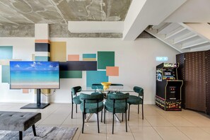 Une chambre moderne dotée d’un grand téléviseur à écran plat, d’une table à manger en verre avec des chaises, d’une console de jeux et d’un plafond en béton.