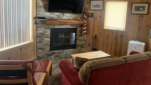 A cozy cabin interior with a stone fireplace, wooden walls, a television mounted above it, and a comfortable seating area with a red sofa and a wooden table.