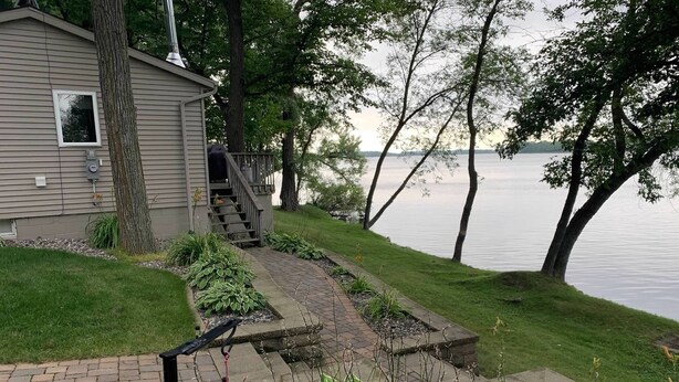 A house with a paved pathway leading to a deck, surrounded by lush greenery and a lake view.