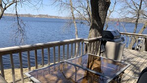 A wooden deck with a glass table and chairs, overlooking a lake.