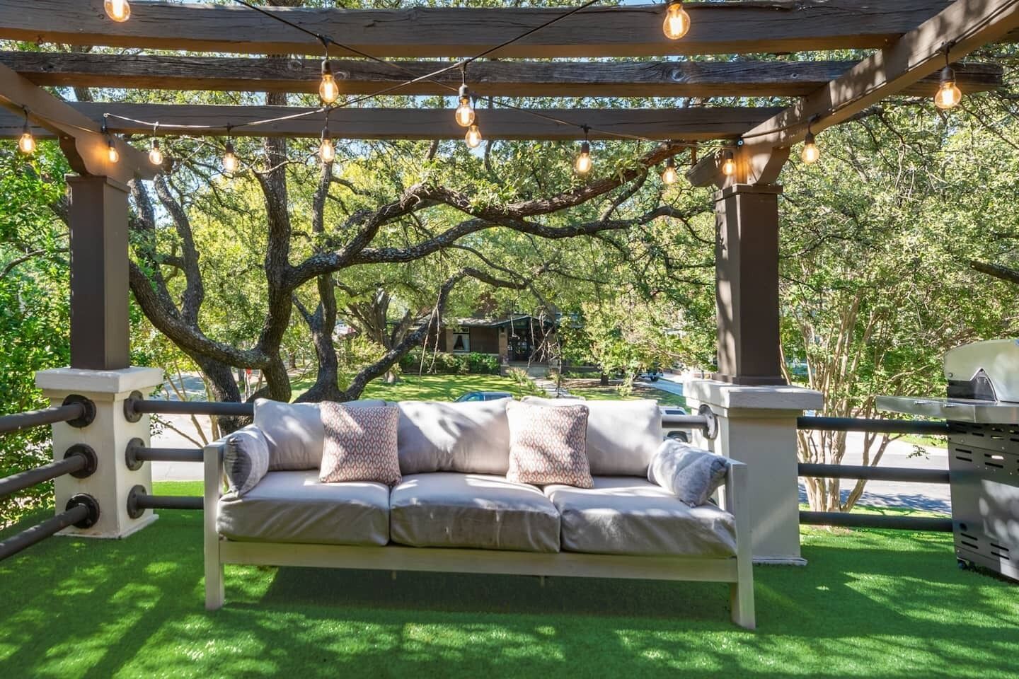 A covered outdoor seating area with a cushioned bench, string lights, and a grill.
