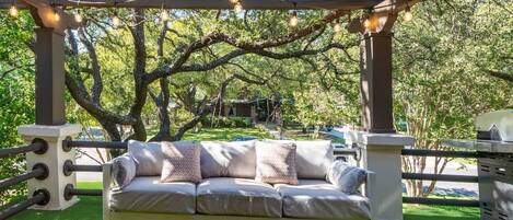 A covered outdoor seating area with a cushioned bench, string lights, and a grill.
