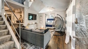 A modern living room with a grey sofa, a hanging chair, a TV mounted on the wall, and a staircase leading to an upper level. - Spacious 2-bedroom loft condo in Dallas’ Historic Munger Place featuring a private designer terrace, secure parking, luxury hybrid mattresses with Egyptian-cotton sheets, and a serene outdoor patio—perfect for relaxing or entertaining. (Dallas)