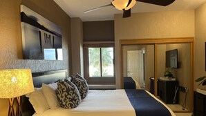 Ein Hotelzimmer mit einem großen Bett, einem Deckenventilator, einem an der Wand befestigten Fernseher und Blick auf Palmen.