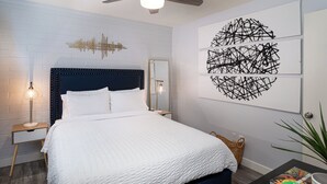 A modern bedroom with a white brick wall, a dark blue headboard, a white quilted bedspread, a ceiling fan, and abstract art on the wall. - Quaint Central Phoenix Duplex   (Phoenix)