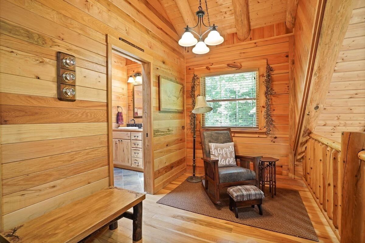 A cozy cabin interior with wooden walls, a bench, a chair with a cushion, a table, and a window with a view of trees.
