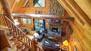 A log cabin interior with a stone fireplace, wooden walls, and a balcony overlooking a deck.