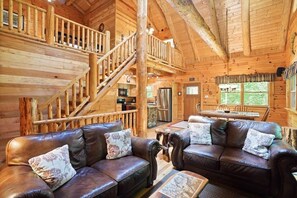 A wooden cabin interior with a staircase, leather sofas, a dining area, and a kitchen.