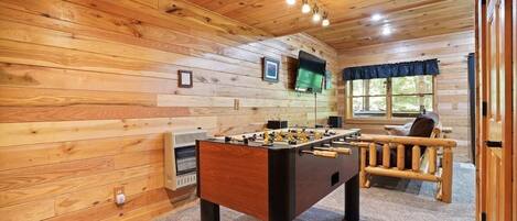 A cozy room with wooden walls, a foosball table, a TV mounted on the wall, and a window with curtains.