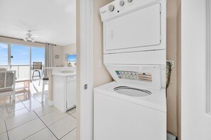 A modern laundry room with a front-loading washer and dryer, a ceiling fan, and a view of a balcony with a chair and table. - 🏖️Beach Front-FREE Beach Service-Pools-Celadon 1103🏖️ (Panama City)