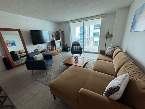 A living room with a sofa, armchairs, a coffee table, a TV, and a large window with a view of buildings. - Gorgeous Apartment in Brickell (Miami)