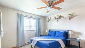 A bedroom with a bed, a ceiling fan, a window with curtains, a bedside table with a lamp, and a shelf with decorative items. - 1BR I Downtown, Queen, Free W/D, Garage (Fargo)