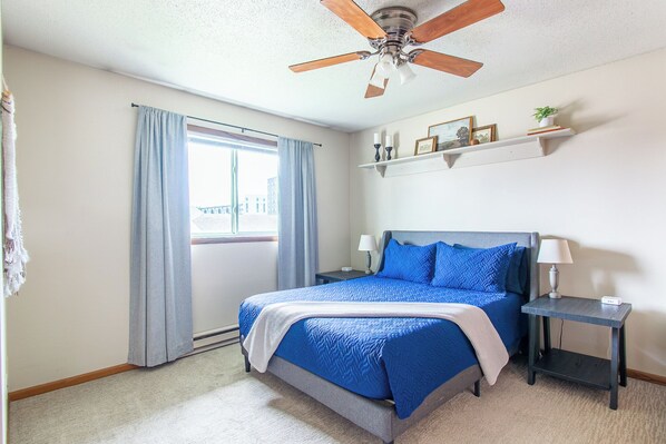 A bedroom with a bed, a ceiling fan, a window with curtains, a bedside table with a lamp, and a shelf with decorative items.