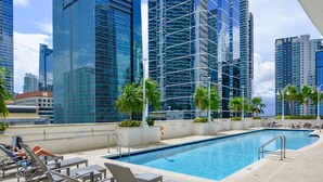 Una piscina sul tetto con poltronne da relax e palme, circondata da grattacieli.
