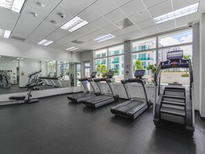 Une salle de sport équipée de tapis de course, dotée d’un mur recouvert de miroirs et de grandes fenêtres offrant une vue sur les bâtiments et les palmiers.