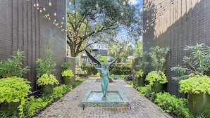 Ein gepflasterter Weg führt zu einem Brunnen mit einer Statue, umgeben von Grünflächen und modernen Gebäuden.