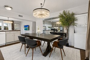 A modern kitchen with a dining area, featuring a chandelier, a large dining table with chairs, and a potted plant. - A spacious two-bedroom condo with an open living area, modern kitchen, large windows, and peaceful views toward the lush courtyard and resort-style pool. (Dallas)