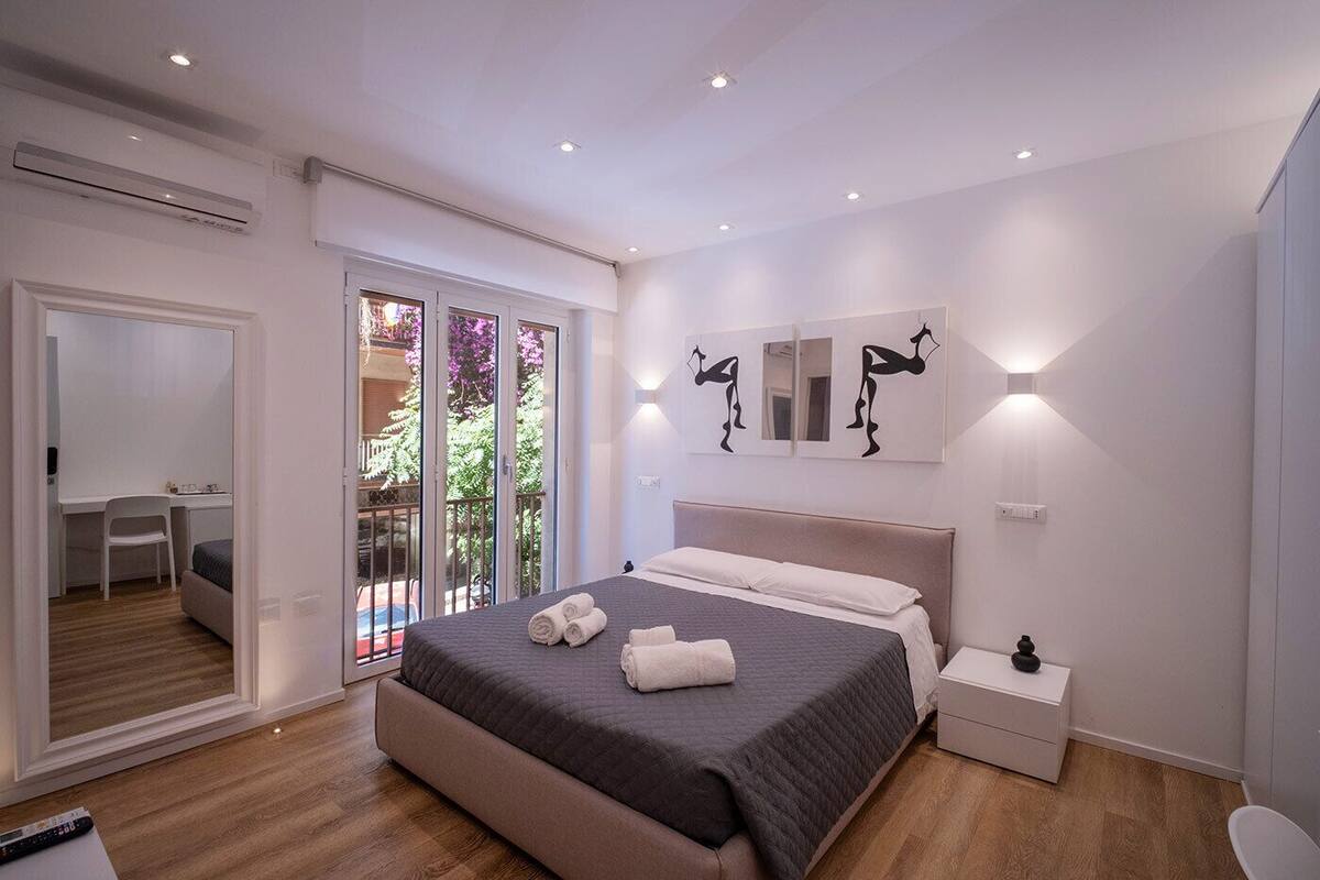 A modern bedroom with a large bed, a desk, and a balcony with a view of trees.