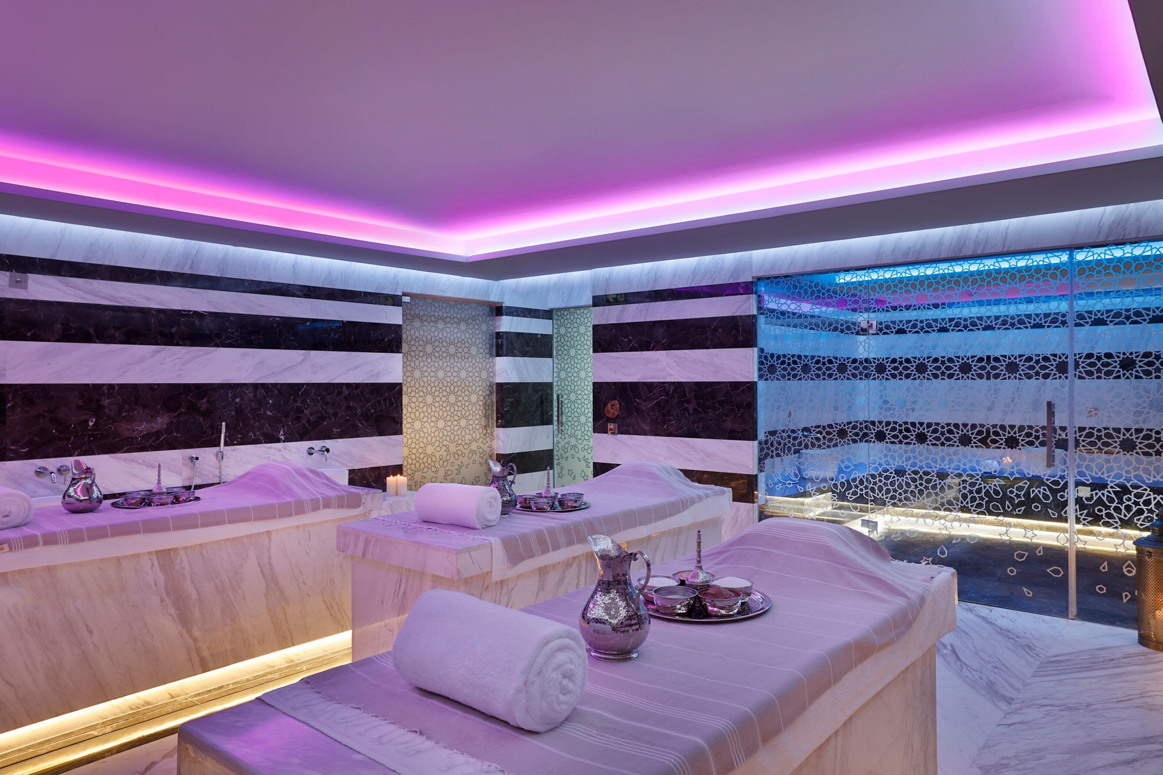 A spa room with two massage tables, each with a towel and a decorative pitcher. The room features a patterned wall and a blue patterned curtain.