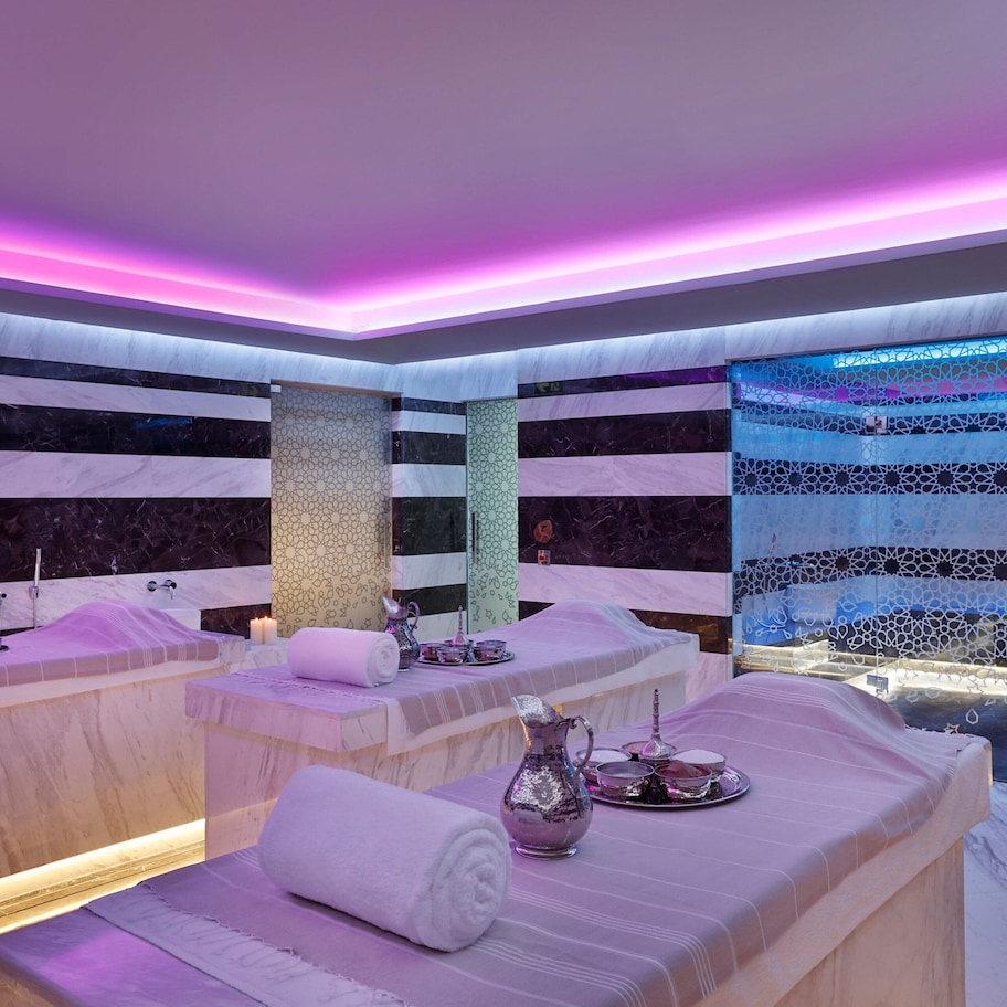 A spa room with two massage tables, each with a towel and a decorative pitcher. The room features a patterned wall and a blue patterned curtain.