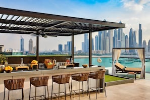 Un bar panoramico sul tetto con vista sullo skyline della città, sgabelli da bar, una zona relax e una piscina.