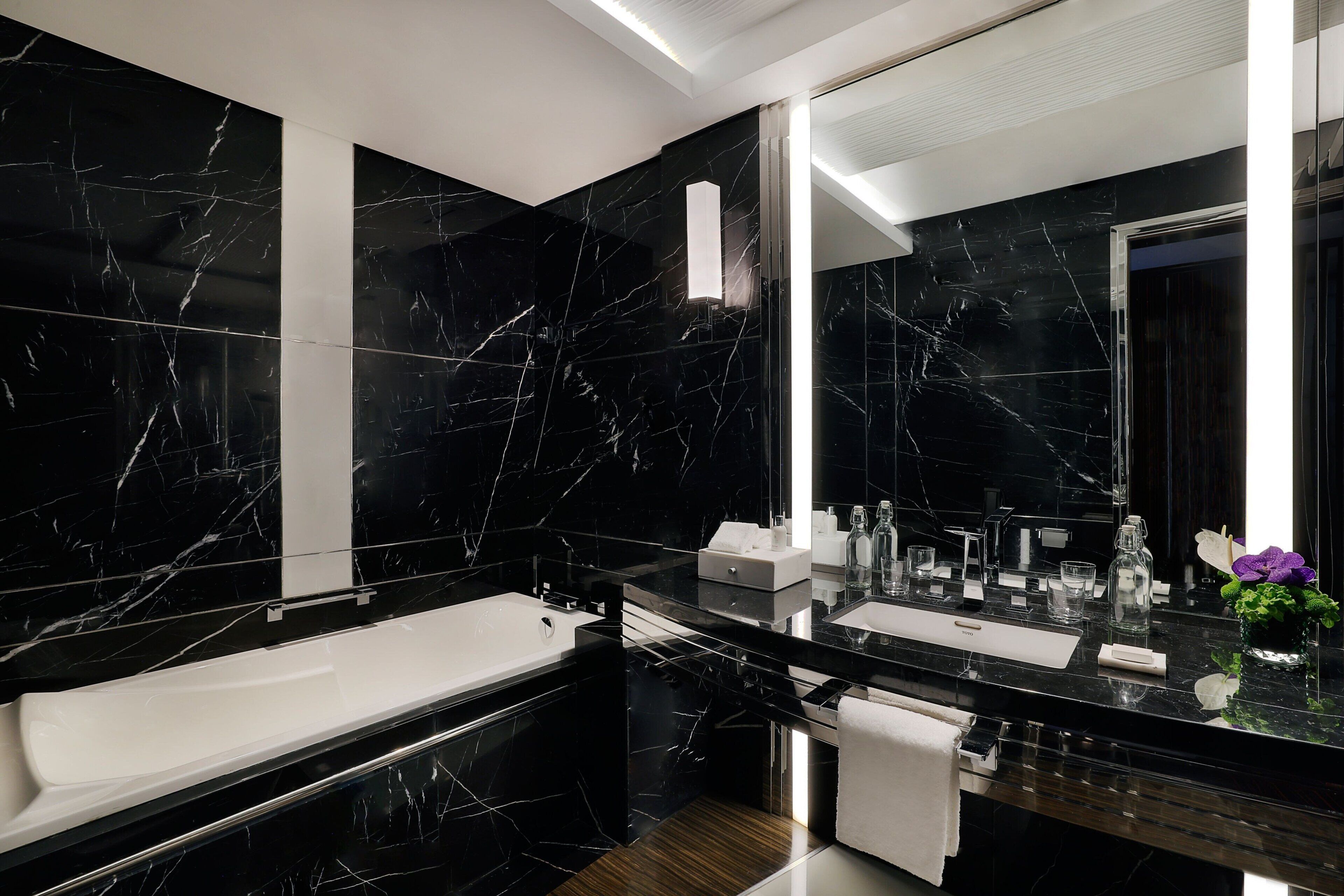 A modern bathroom with black marble walls, a large mirror, a white bathtub, and a wooden vanity.