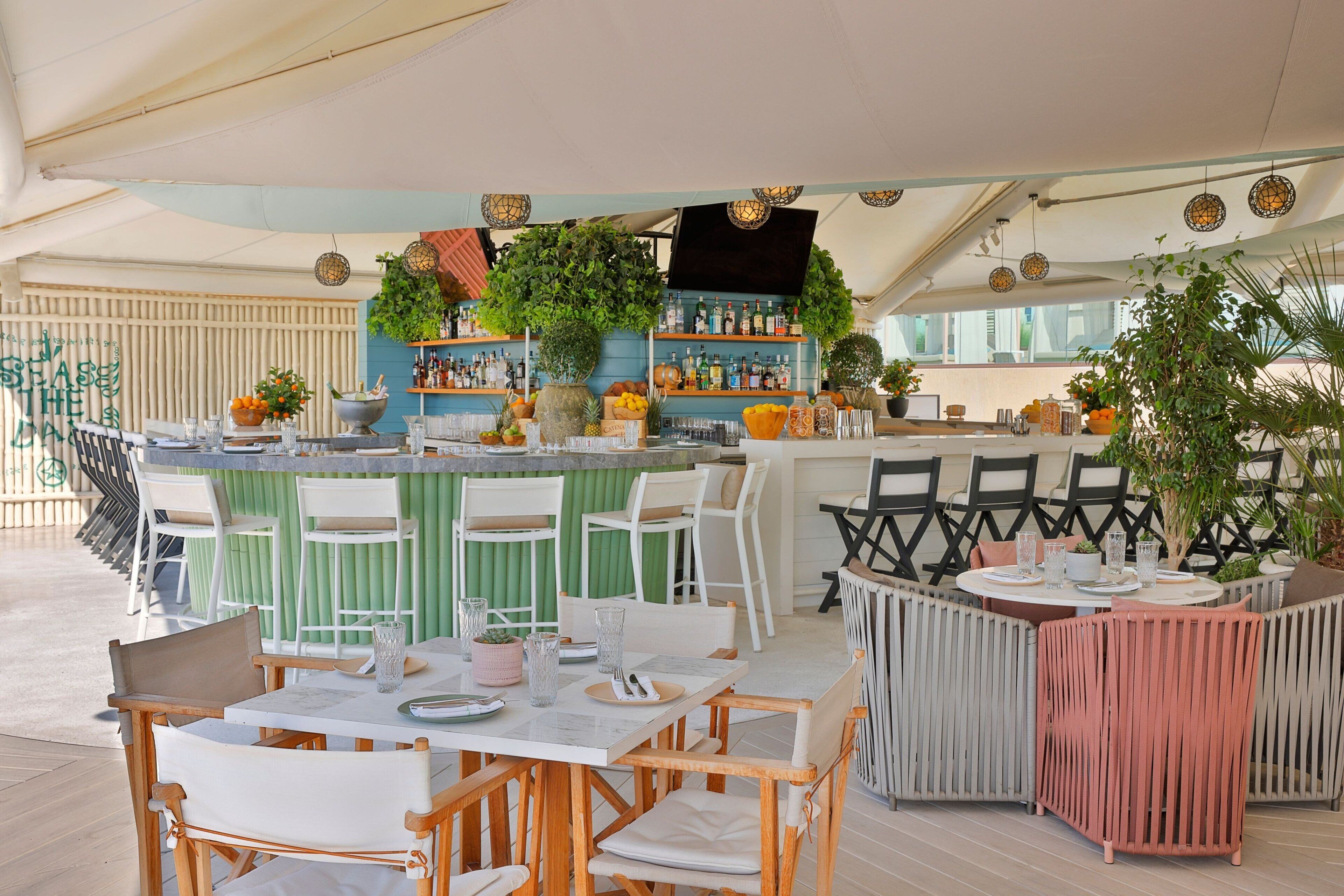 An outdoor dining area with multiple tables and chairs, a bar with shelves of bottles, and potted plants.