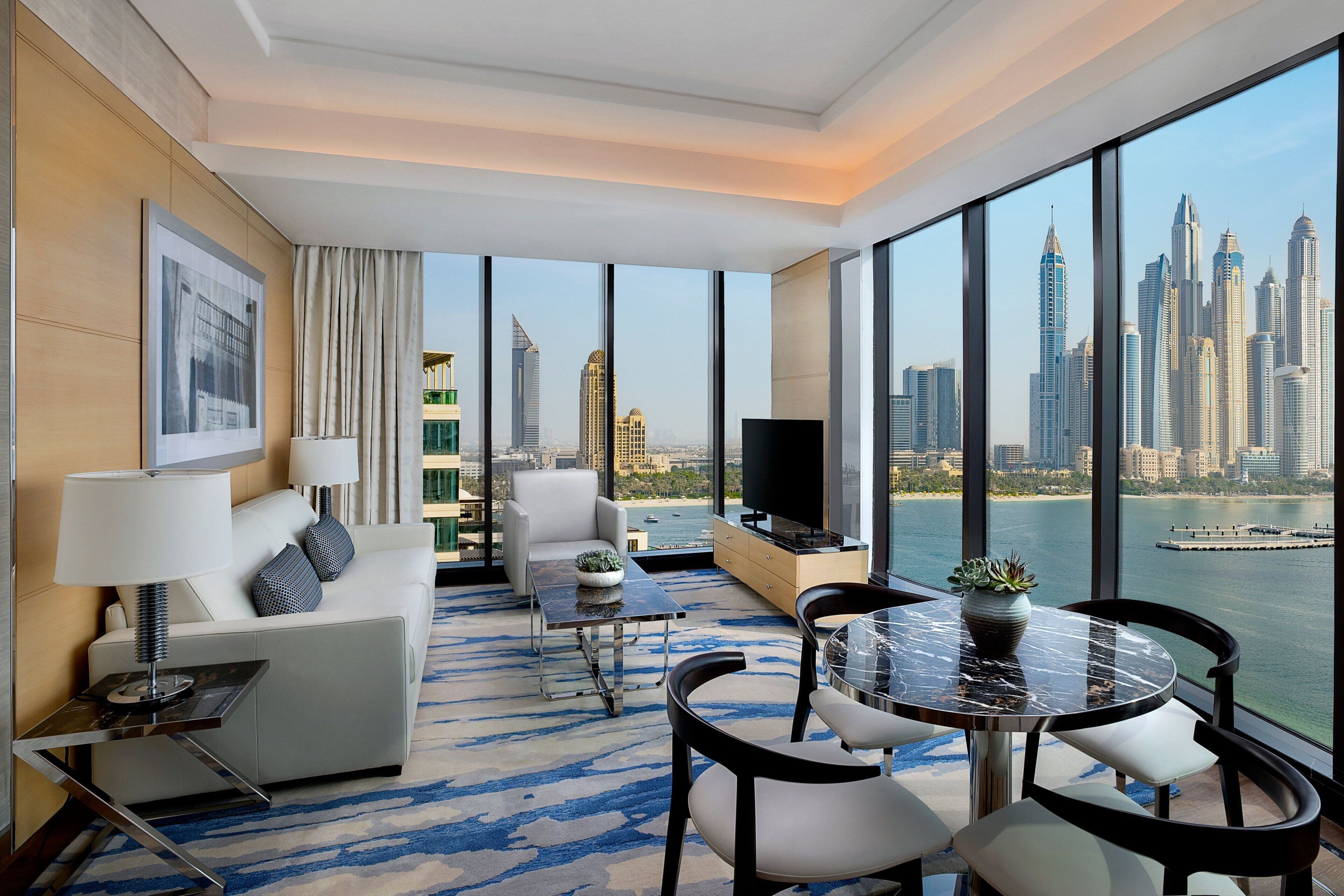A modern hotel room with a large window offering a city skyline view, a sofa, a dining table, and a TV.