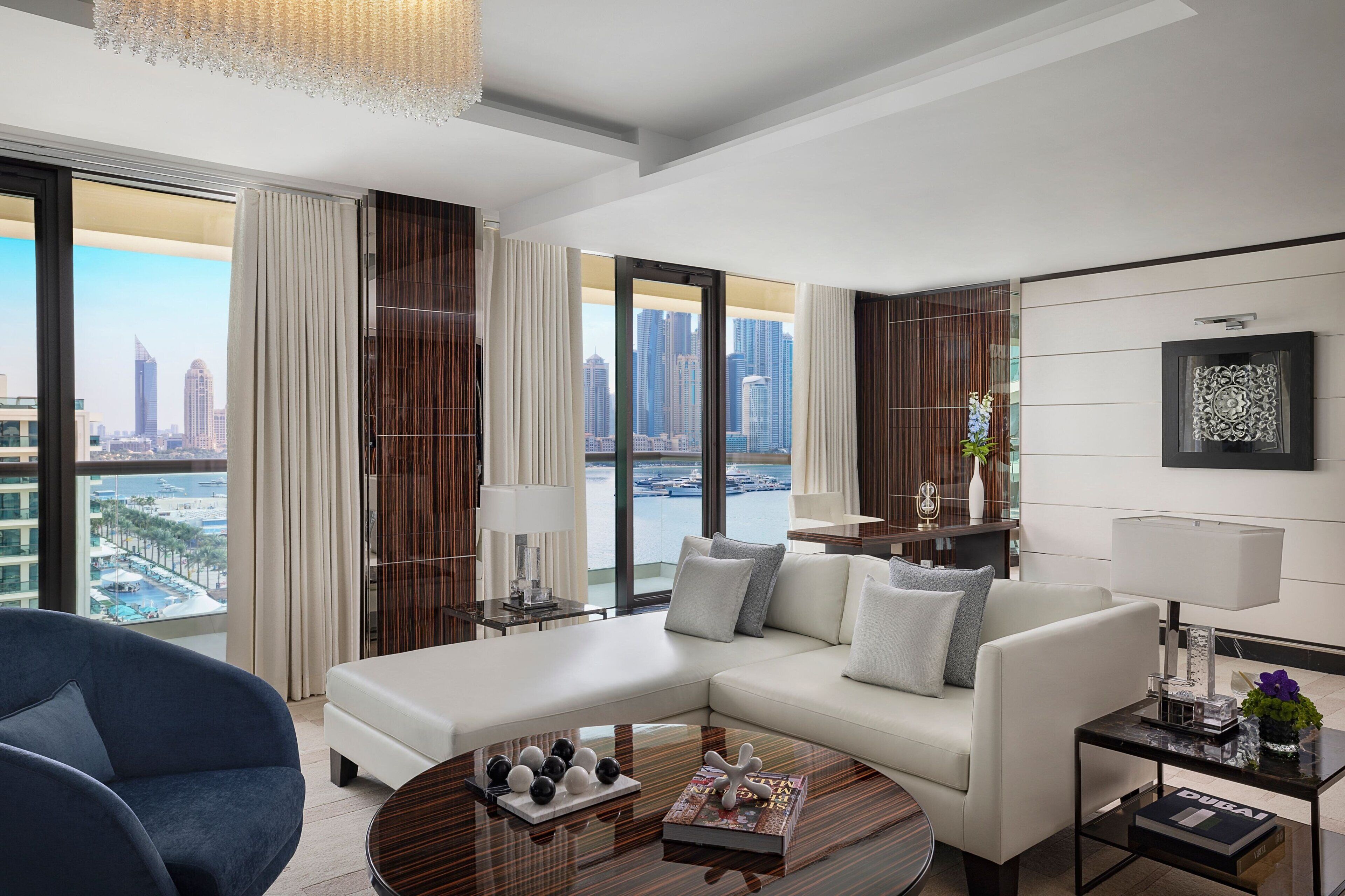 A modern living room with a white sofa, a blue armchair, and a coffee table with decorative items. Large windows offer a view of a city skyline and a body of water.
