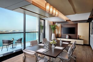 A modern conference room with a large table, chairs, and a TV. It has a view of the ocean and a balcony with chairs.