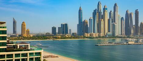 A coastal cityscape with modern high-rise buildings, a beach with sun loungers, and a marina.
