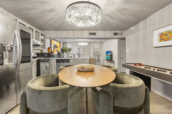 A modern kitchen with a round dining table, grey upholstered chairs, stainless steel appliances, and a chandelier.