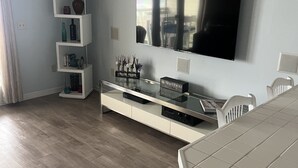 A modern living room with a flat-screen TV mounted on the wall, a white entertainment unit with electronic devices, a dining table with chairs, and a wooden floor.