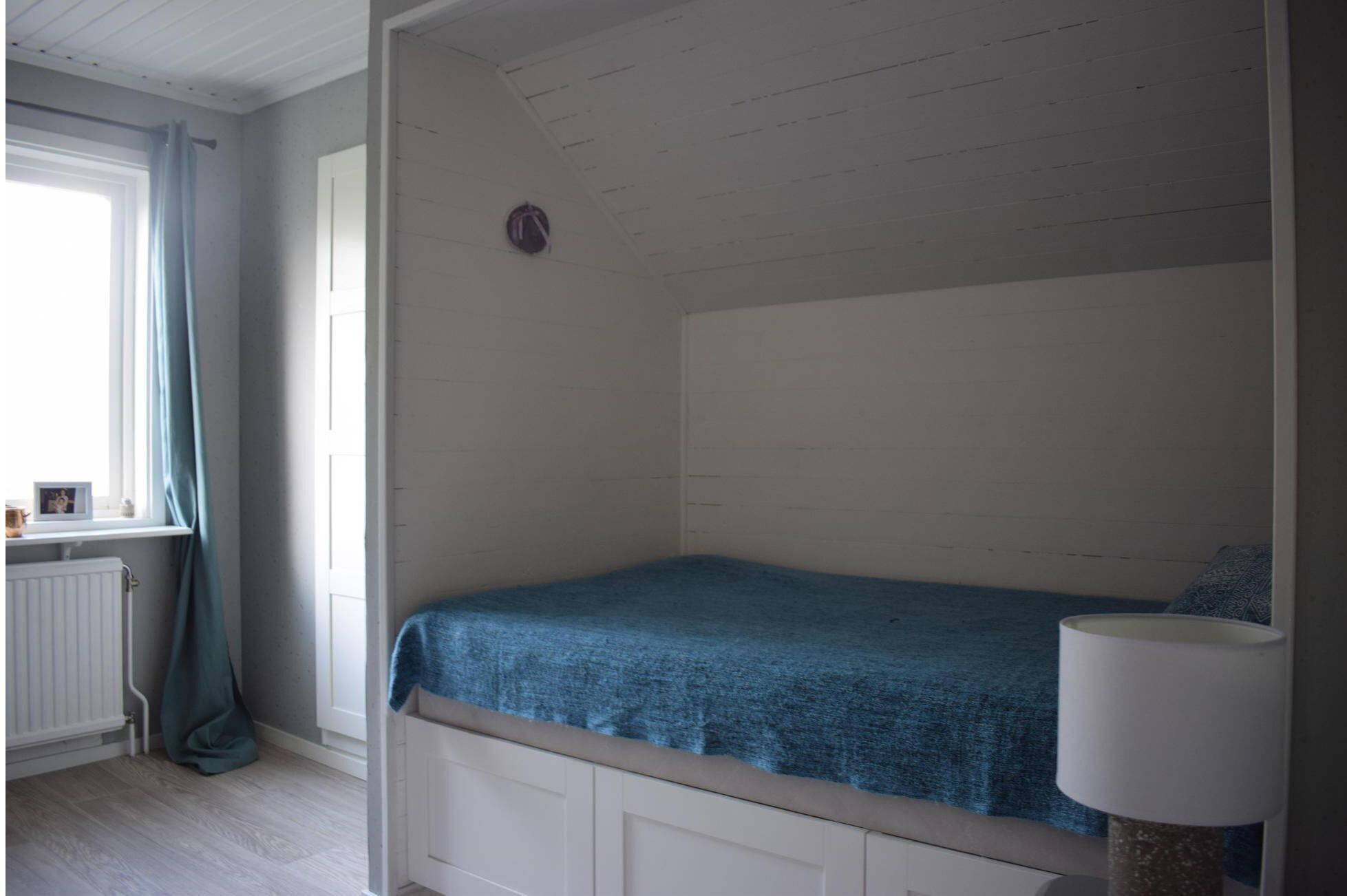 Una cama individual con colcha azul en una habitación con paredes blancas y una ventana con cortinas.