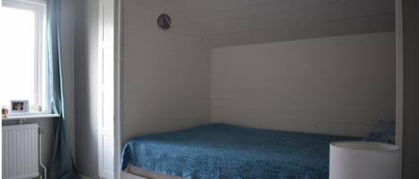Una cama individual con colcha azul en una habitación con paredes blancas y una ventana con cortinas.