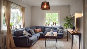 A modern living room with a dark blue sectional sofa, a wooden coffee table, and a hanging pendant light. There are potted plants on the windowsill and a flat-screen TV on a wooden stand. - Awesome home in Kalmar with WiFi (Virserum)