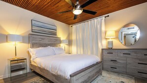 Een slaapkamer met een houten bed, een plafondventilator, een nachtkastje met een lamp, een dressoir en een spiegel aan de muur.