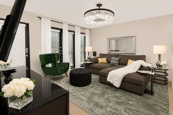 A modern living room with a sectional sofa, a green armchair, a black ottoman, a glass coffee table, and a chandelier. - An inviting two-bedroom condo with a bright open layout, tranquil garden views, and a private patio overlooking a beautifully landscaped courtyard and pool.🎡 STAY MINUTES FROM THE STATE FAIR OF TEXAS! 🎡 (Dallas)