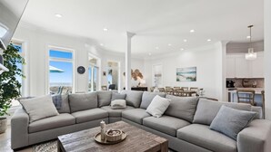 A modern living room with a large sectional sofa, a wooden coffee table, and a dining area with a view of the ocean.