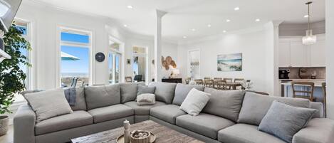 A modern living room with a large sectional sofa, a wooden coffee table, and a dining area with a view of the ocean.