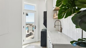 A modern kitchen with a white countertop, a black appliance, and a view of a pool and beach through a glass door.