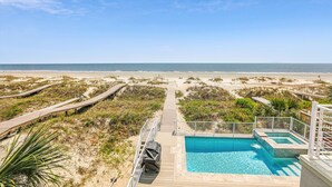 A beachside pool area with a wooden deck leading to the pool, surrounded by sand dunes and vegetation.
