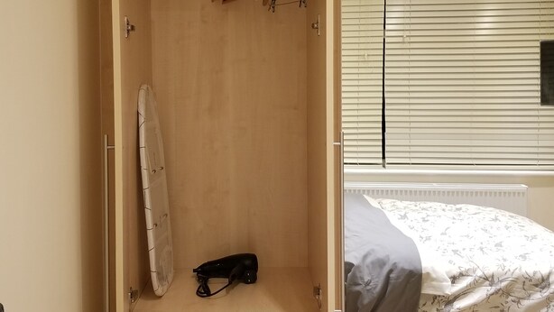 A bedroom with a wooden wardrobe, a bed with a floral bedspread, and a window with blinds. - London Luxury 1 bed studio 5 mins from Ilford Stn free parking (London)
