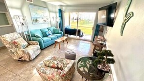 A living room with a blue sofa, floral armchairs, a glass coffee table, and a TV mounted on the wall. - BEACHFRONT Ground Floor WALK-OUT 3Bdrm/2Ba Plantation Palms Great for Families (Gulf Shores)