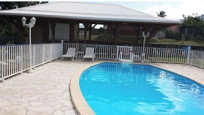 A swimming pool with a tiled deck, a covered seating area with chairs, and a fenced perimeter. - Spacious T3 gîtes, lovely terrace and shared swimming pool, sea view, peaceful (Capesterre-Belle-Eau)