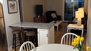 Una cocina moderna con encimera de granito, armarios blancos y un refrigerador pequeño. Cuenta con un comedor con mesa y sillas blancas, y una sala de estar con sofá negro y una lámpara.
