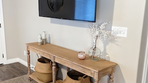 A wooden console table with woven baskets underneath, a flat-screen TV mounted on the wall, and a small decorative vase with branches.