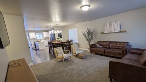 A spacious living room with a brown leather couch, a dining table with chairs, and a kitchen in the background.